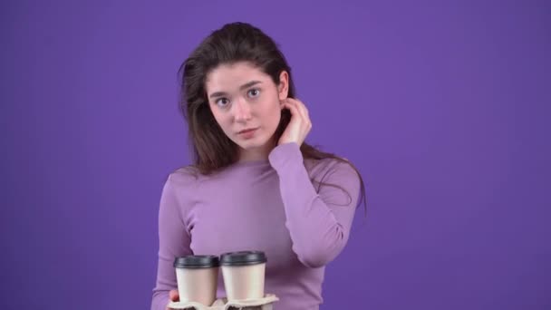 Le portrait d'une fille charismatique porte honteusement ses cheveux derrière ses oreilles, avec quatre verres de thé qu'elle répand devant elle. Brunette isolée sur fond violet, vêtue d'un violet — Video