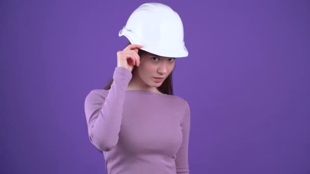 The mysterious young woman in the white helmet of the engineer looks up with her hand on her cap, then punches her with both fists, displays a wide smile. Brunette isolated on a purple background — Stock Video