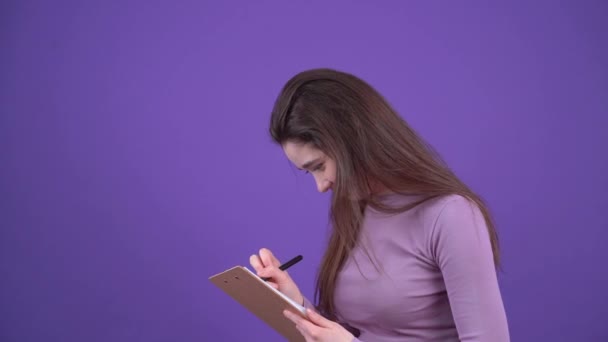 A jovem focada toma notas e planeja algo, escreve ideias, olha pensativamente e escreve. Morena isolada em um fundo roxo, vestida com uma camisola roxa. Conceito de estilo de vida — Vídeo de Stock