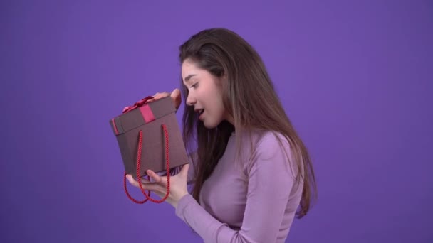 A jovem curiosa abre o presente e parece chocada, exibindo um sorriso largo. Morena isolada em um fundo roxo, vestida com uma camisola roxa. Conceito de estilo de vida — Vídeo de Stock