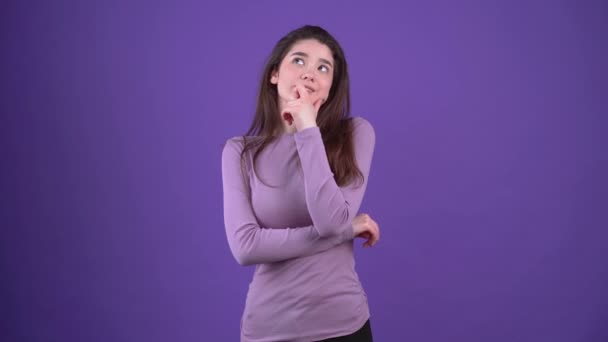 The beautiful, serious girl is sitting with her hand on her chin and thinking about the solution. Brunette isolated on a purple background, dressed in a purple sweater. Lifestyle concept — Stock Video