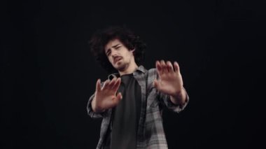 The curly, charismatic boy stretches out his palms forward and frowns and says no, leaning back. Isolated on black background, Concept of life. Peoples emotions. 4k portrait