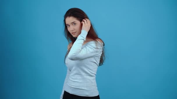 El retrato de una joven tímida que se gira hacia un lado, sonríe coqueta y se pone el pelo detrás de la oreja. Asiático con el pelo oscuro, vestido con una blusa azul, aislado sobre un fondo azul oscuro en el — Vídeos de Stock