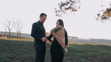 Birbirine aşık genç insanlar el ele tutuşuyor ve mutlu bir şekilde gülümsüyor, büyük bir buket çiçek tutuyor ve teşekkür etmek için onu öpüyor. Doğanın ortasında, gün batımında, yalnız bir çift.