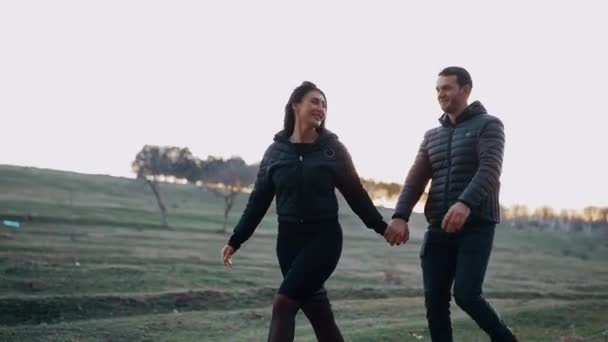 De verliefde jongeren schudden elkaar de hand, de vrouw springt naar voren en de man volgt haar, verbaasd over haar energie, midden in de natuur, onder het prachtige licht van de zonsondergang. Levensstijl concept van — Stockvideo