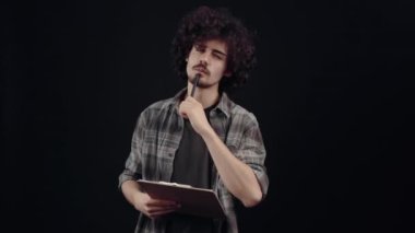 Kıvırcık, ciddi, düşünceli genç adam bir eylem planı hazırlıyor ve bunları ajandasına yazıyor. Siyah arka planda izole edilmiş, yaşam kavramı. İnsanların duyguları işte. 4k portre