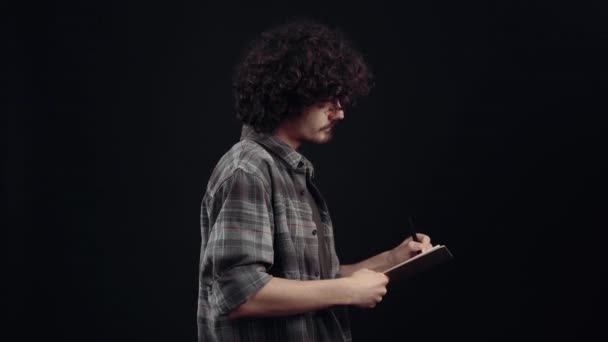 Привлекательный мальчик с вьющимися волосами, в профиль, думает, пишет, отмечает с ручкой на простыне. Изолированный на черном фоне, Концепция жизни. Народные эмоции. Портрет 4k — стоковое видео