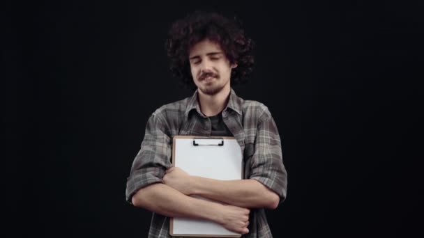 O menino bonito, hipster com cabelo encaracolado, exibe um sorriso largo, segura um planejador e uma caneta em seus braços. Isolado em fundo preto, conceito de vida. Emoções dos povos. Retrato 4k — Vídeo de Stock