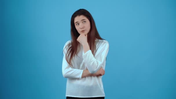 The young woman, thinking with her hand on her chin, looks in parts and thinks how to proceed, what to choose. Asian with dark hair, dressed in a blue blouse, isolated on a dark blue background in the — Stock Video