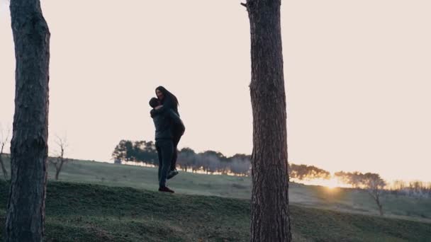 Os jovens apaixonados, vestidos de preto, abraçam-na firmemente, o homem pega-lhe nos braços e levanta-a com muito amor, a mulher sorri de largura, feliz, no meio da natureza à luz do pôr do sol — Vídeo de Stock