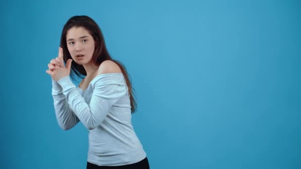 De jonge agent simuleert een pistool, spioneert, simuleert een schot en blaast rook uit het pistool. Aziatisch met donker haar, gekleed in een blauwe blouse, geïsoleerd op een donkerblauwe achtergrond in de studio. De — Stockvideo