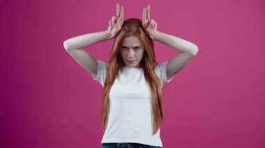 Sinirli genç kadın ellerini boynuz şeklinde başına koyar ve ileride hayal kırıklığına uğramış gibi görünür. Çilli genç, beyaz tişörtlü, pembe arka planda izole edilmiş. İnsanların konsepti