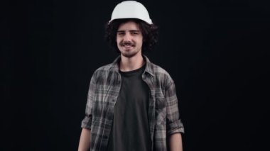 Kafasında beyaz mühendis kaskı olan karizmatik hippi kaşlarıyla flört ediyor ve gururla ileriye bakıyor. Siyah arka planda izole edilmiş. Hayat kavramı. İnsanların duyguları işte. 4k portre