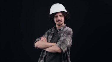 Kafasında mühendis kaskı olan yakışıklı genç adam ellerini göğsünde çapraz yapar ve gururla kaşlarını kaldırır. Siyah arka planda izole edilmiş. Hayat kavramı. İnsanların duyguları.