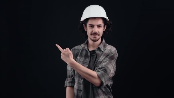 El carismático hipster con el casco blanco de los ingenieros se ve alegre con su dedo índice a un lado. Aislado sobre fondo negro. El concepto de vida. Emociones de la gente. Retrato 4k — Vídeos de Stock