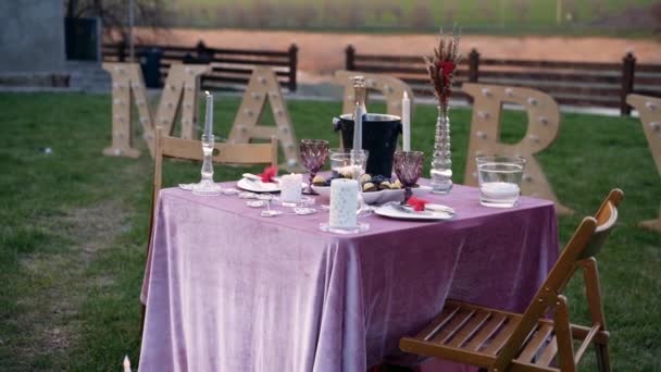 Der romantische Rahmen, von der grünen Wiese, unter dem rustikalen Hintergrund des schön dekorierten Tisches, mit Kerzen, Besteck, Bonbons und Champagner. Dahinter stehen Großbuchstaben in Form von — Stockvideo