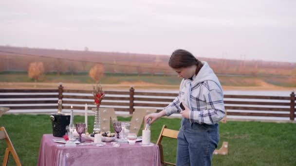 Das romantische Dekor im Freien vor dem Hintergrund einer rustikalen Aussicht vorbereitet. Ein Tisch mit Gläsern, Champagner, Pralinen, Kerzen, dekorativen Blumen. Das Mädchen zündet die Kerze mit einem anderen an — Stockvideo