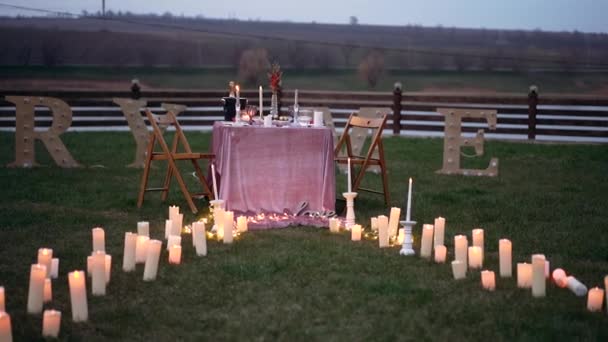Romantisk kvällsmiljö, ljusen som bildar en stig, vid vars ändar det finns ett bord dekorerat med ljus, champagne, blommor och snacks. versaler placerade på gräset nedanför texten — Stockvideo