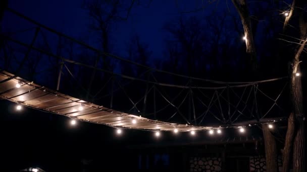 Il paesaggio di un ponte di legno illuminato nel buio della notte lampadina, circondato dalle silhouette degli alberi. Ambiente romantico, atmosfera rilassante. Ritratto 4k — Video Stock