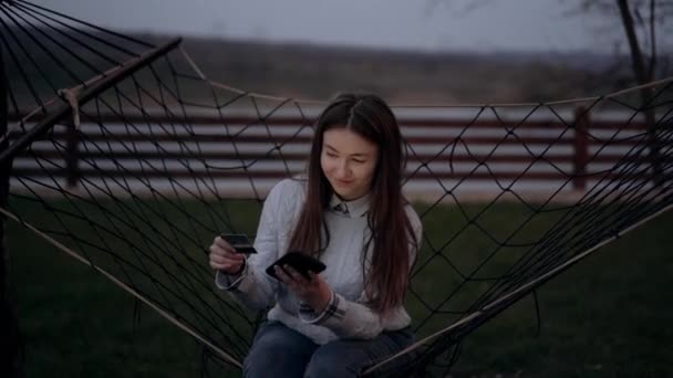 La joven hermosa mujer hace compras en línea con su tarjeta y teléfono en un columpio fuera, en el contexto de la naturaleza verde, en el ambiente de una noche tranquila. Retrato 4k — Vídeos de Stock