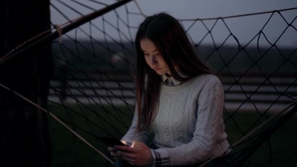Chica concentrada vestida con suéter azul, en el silencio de la noche, sentada en el columpio exterior, escribiendo enfocada en la pantalla del teléfono, atrapada en la conversación. Paisaje nocturno en la naturaleza. 4k — Vídeos de Stock