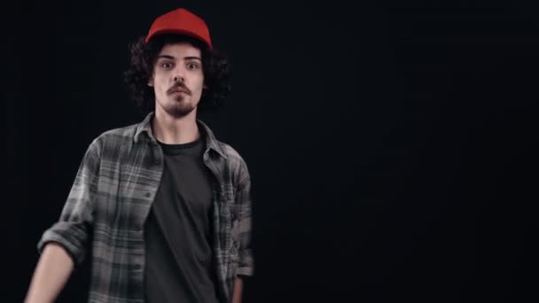 Le portrait d'un hipster optimiste avec une casquette rouge pointant son index sur un côté montre alors le même signe. Isolé sur fond noir. Le concept de vie. Des émotions populaires. 4k — Video