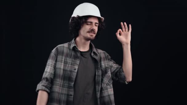 El joven ingeniero, optimista, con un casco blanco, muestra el signo de ok con sus dedos que conduce a ok a continuación, a través de las partes. Aislado sobre fondo negro. El concepto de vida. Pueblos — Vídeos de Stock