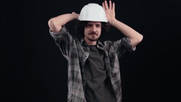 这位幽默的工程师头戴白色头盔，乐观地把手掌放在头上，做成兔子耳朵的形状，并玩弄它们。被黑色背景隔离。生命的概念。人民 — 图库视频影像