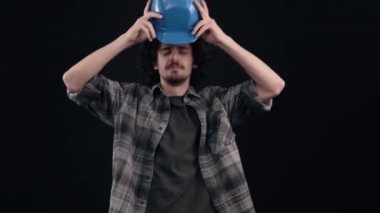 Genç mühendis, mutlu, karizmatik, mavi kaskıyla dans ediyor. Siyah arka planda izole edilmiş. Hayat kavramı. İnsanların duyguları işte. 4k portre