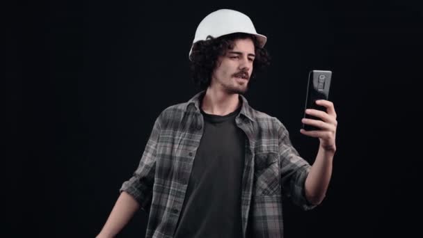기술자와 함께 헬멧을 쓴 젊은이는 비디오를 통해 전화로 이야기하고 악수를 한다. 검은 배경에 고립되어 있습니다. 생명의 개념. 국민의 감정. 4k 사진 — 비디오