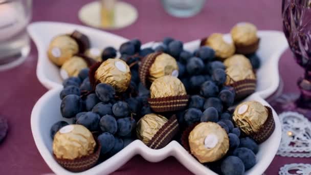 Placa de mariposa blanca llena de chocolates bien colocados en la uva. Pequeño detalle en la mesa donde tiene lugar la cena romántica. Retrato 4k — Vídeos de Stock