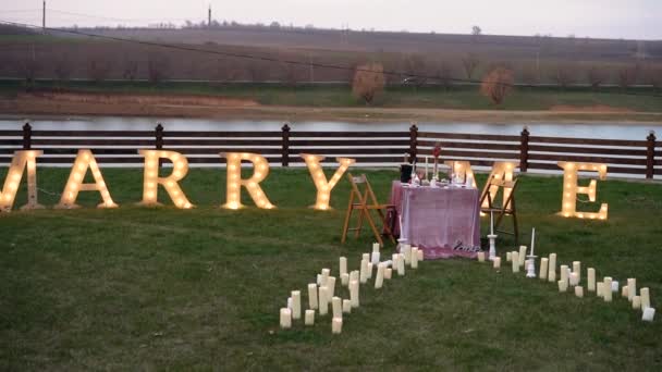 O cenário romântico de uma proposta de casamento, uma mesa decorada com flores, talheres, velas e champanhe, em um prado verde à beira de um lago. Letras maiúsculas colocadas na grama abaixo do texto — Vídeo de Stock
