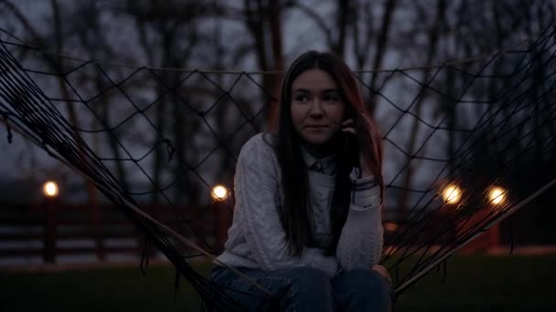 Kvällsporträtt av en flicka med en grå tröja och långt brunt hår sitter på en gunga och pratar allvarligt på telefonen. Nattlandskap i naturen med träd och kvällslampor. 4k porträtt — Stockvideo