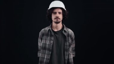 Makinistin beyaz kasklı portresi kameraya bakıyor ve geniş bir gülümseme sergiliyor, şık kaşlar yapıyor. Siyah arka planda izole edilmiş. Hayat kavramı. İnsanların duyguları işte. 4k