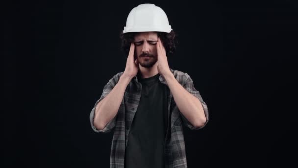 L'ingénieur débutant avec un casque blanc, après une longue journée de travail, souffre de migraines, tient ses tempes des deux mains. Isolé sur fond noir. Le concept de vie. Des émotions populaires. 4k — Video