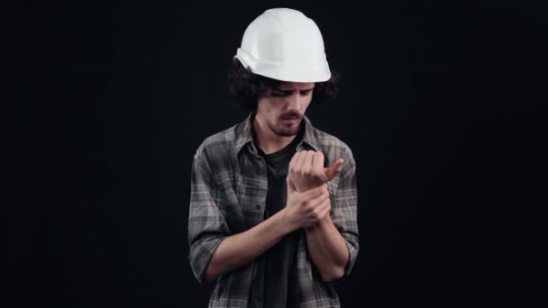 Инженерный мальчик в белом шлеме, к сожалению, цепляется за запястье, страдает от сильной боли. Изолированный на черном фоне. Концепция жизни. Народные эмоции. Портрет 4k — стоковое видео
