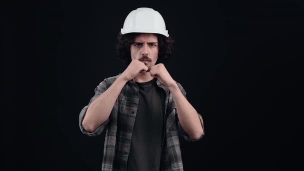 El ingeniero enojado, con un casco blanco, está listo para luchar, levanta los puños nerviosamente en una posición de lucha. Aislado sobre fondo negro. El concepto de vida. Emociones de la gente. Retrato 4k — Vídeos de Stock