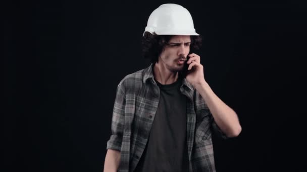 The engineer with the white helmet speaks on the phone, finds out bad news and angrily shouts back with a loud gesture of his hand. Isolated on black background. The concept of life. Peoples emotions — Stock Video