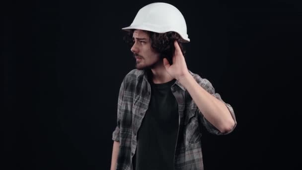 El joven ingeniero, con un casco blanco, se pone la mano en la oreja, no oye lo que se le dice, cuando oye, frunce el ceño y dice que no. Aislado sobre fondo negro. El concepto de vida — Vídeos de Stock