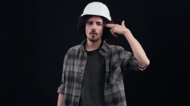 Mühendis miğferindeki karizmatik genç adam parmaklarını şakağa tabanca şeklinde sokar. Siyah arka planda izole edilmiş. Hayat kavramı. İnsanların duyguları işte. 4k portre