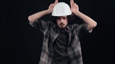Mühendis miğferindeki karizmatik çocuk parmaklarını öfkeyle boynuz şeklinde kafasına dayar. Siyah arka planda izole edilmiş. Hayat kavramı. İnsanların duyguları işte. 4k portre