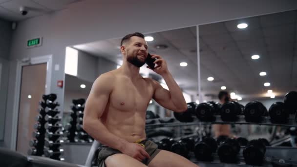 De jonge sporter, in de sportschool, neemt een pauze en praat aan de telefoon met een vriend, glimlacht breed en verheugt zich tijdens de pauze tussen de oefeningen. Intense mannelijke energie. Een gezonde levensstijl. Sport — Stockvideo