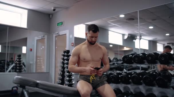 El joven deportista, centrado, durante el descanso entre ejercicios escribe un mensaje por teléfono. Intensa energía masculina. Estilo de vida saludable. Concepto deportivo — Vídeos de Stock