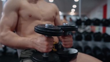 Spor sandalyesinde oturan kaslı sporcunun elindeki dambıllara odaklan. Yoğun erkeksi enerji. Sağlıklı bir yaşam tarzı. Spor konsepti