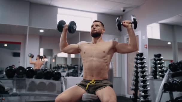 El hombre enorme y deportivo realiza levantar las mancuernas, en el gimnasio, en una posición sentada. Intensa energía masculina. Estilo de vida saludable. Concepto deportivo — Vídeo de stock