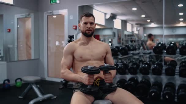 De knappe, gespierde man zit in de sportschool, met twee halters in zijn hand, hard ademend en voorover leunend. Intense mannelijke energie. Een gezonde levensstijl. Sport concept — Stockvideo