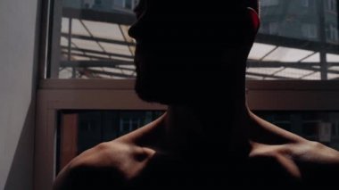 Confident young man standing by the window, muscular with bare chest, silhouette in semi-darkness. Intense masculine energy. Healthy lifestyle. Sport concept