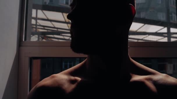 Confident young man standing by the window, muscular with bare chest, silhouette in semi-darkness. Intense masculine energy. Healthy lifestyle. Sport concept — Vídeos de Stock