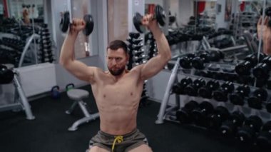 Genç koç, ellerini oturma pozisyonundan nasıl doğru bir şekilde dambılları kaldıracağını gösteriyor. Yoğun erkeksi enerji. Sağlıklı bir yaşam tarzı. Spor konsepti