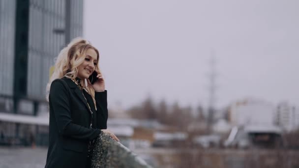 De blonde zakenvrouw praat in een goed humeur aan de telefoon, glimlacht breed, tegen de achtergrond van de luidruchtige stad, gekleed in een jas, met een schelle make-up. Vrouwelijke energie. Schoonheidssector — Stockvideo